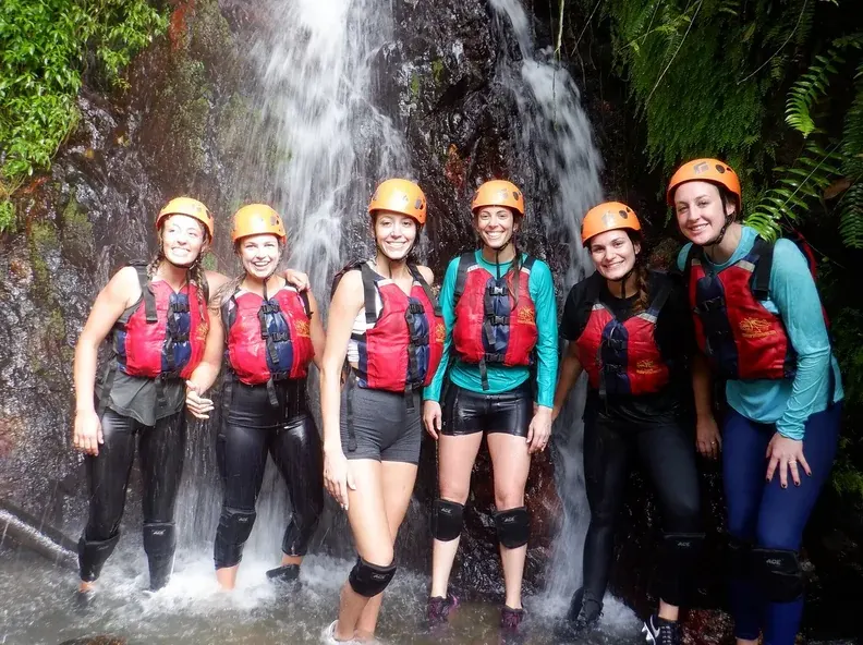 Group of six guests in helmets and life jackets standing under a rainforest waterfall
