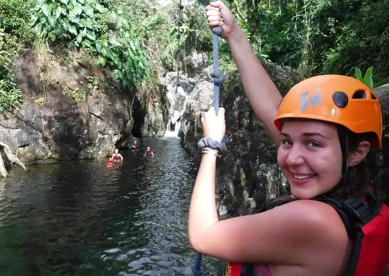 Guest in a helmet and life jacket gripping the rope swing above a rainforest pool